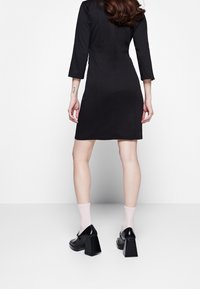 Musta vartalonmyötäinen mekko, jossa on kolme neljäsosahihaa ja sileä kangas. Yhdistetty vaaleanpunaisiin sukkiin ja mustiin lohkopohjaisiin korkokenkiin. Yksinkertainen, moderni design.