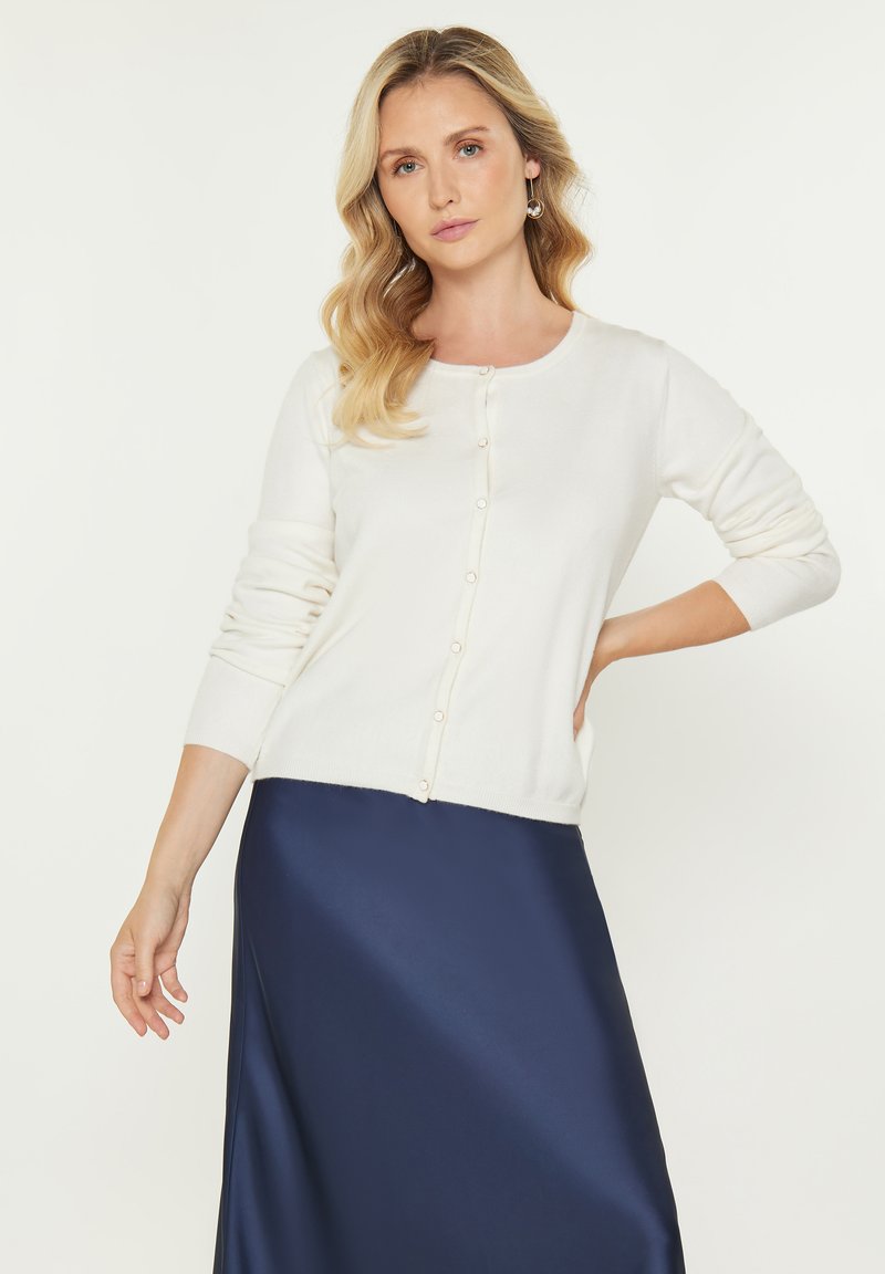 Witte gebreide cardi met lange mouwen, ronde halslijn en parelknopen, gecombineerd met een donkerblauwe satijnen rok, gepresenteerd op een lichte achtergrond.