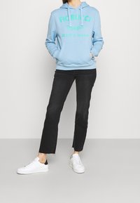 Sudadera con capucha azul claro con texto gráfico verde, bolsillo frontal y cordones. Combinada con vaqueros negros y zapatillas blancas con detalles negros.