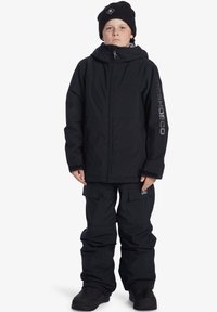 Svart snöjacka med huva, dragkedja fram och logotyp på ärmen, ihop med matchande cargobyxor och svarta stövlar. Slät, vattentålig yta.