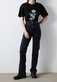 Černé bavlněné tričko s třpytivým grafikou růže a bílým textem, spárované s černými zvonovými džíny s vysokým pasem a černými botami.
