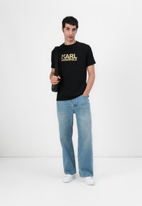 Camiseta de algodón negra con el logo dorado "KARL LAGERFELD", combinada con jeans de pierna ancha de color azul claro y zapatillas blancas. Diseño simple, ajuste relajado.