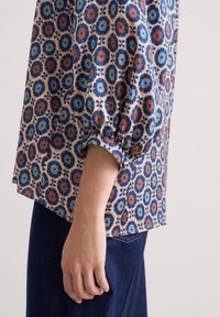 Die Bluse mit Blumenmuster besteht aus einem weichen, leichten Stoff in Blau-, Rot- und Beigetönen, hat geraffte Ärmel und eine lockere Form.