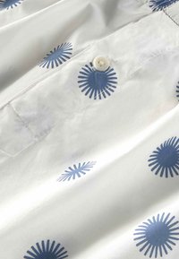 Weißer Stoff mit blauen Sonnenstrahlenmustern; verfügt über einen Knopf und sichtbare Nähte, zeigt eine glatte Textur und ein strukturiertes Design.