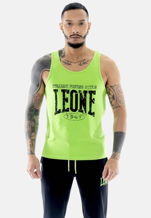 Homme avec des tatouages portant un débardeur vert vif avec le texte "LEONE 1947" et un pantalon noir, debout devant un fond blanc uni.