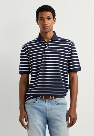 Junger Mann trägt ein marineblau-weiß horizontal gestreiftes Poloshirt, das in eine hellblaue Jeans mit braunem Gürtel gesteckt ist, und steht vor einem einfarbigen Hintergrund.