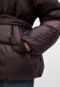 Smeđa podstavljena jakna s pojasom u struku, kviltanom teksturom i širokim rukavima. Ima bočne džepove s glatkom završnom obradom.