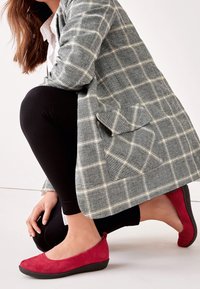 Femme à genoux en leggings noirs, blazer gris à carreaux avec poche à rabat, chemise blanche et chaussures plates rouges avec semelles noires sur un sol blanc.