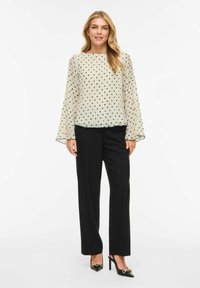 Przezroczysta kremowa bluzka w czarne grochy, z długimi rozkloszowanymi rękawami, zestawiona z czarnymi spodniami i czółenkami na szpic. Obecne delikatne detale teksturalne.