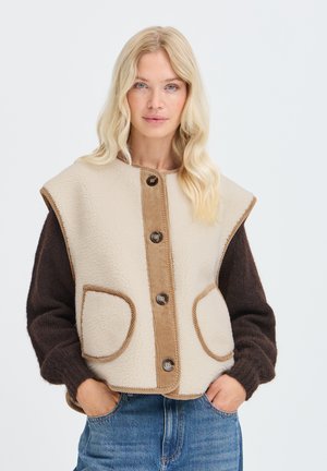 Donna bionda che indossa un gilet in pile beige con bottoni sopra una maglia a maniche lunghe marrone scuro, con le mani nelle tasche dei jeans blu.