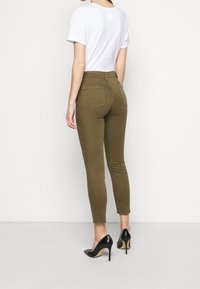 Jeans skinny verde oliva hechos de mezcla de algodón, con bolsillos traseros y cintura de altura media, combinados con una camiseta blanca y tacones negros.