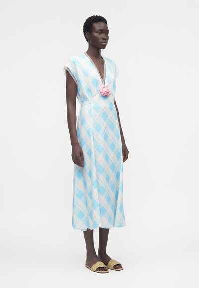 Γυναίκα που φοράει μπλε-λευκό μίντι φόρεμα με μοτίβο σε σχήμα ρόμβου και δαντελένιο τελείωμα, με ροζ καρφίτσα λουλούδι, συνδυασμένο με μπεζ σανδάλια τύπου slide.