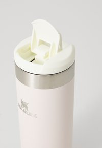 Tasse de voyage en acier inoxydable avec une finition mate rose pâle, dotée d'un couvercle rabattable blanc et d'une bande d'accent argentée autour du haut.