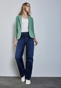 Muntgroene blazer, witte tanktop en donkerblauwe wijde jeans. Witte sneakers maken de outfit af, met een soepele stoftextuur en een nauwkeurige snit.