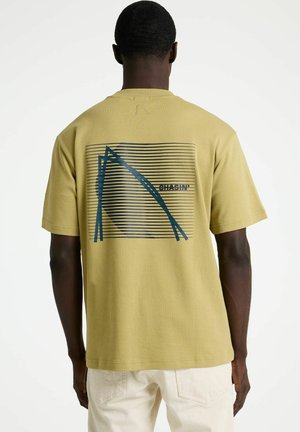 Man draagt een mosterdgele t-shirt met een abstract blauw-zwart gestreept grafisch ontwerp en de tekst "CHASIN'" op de achterkant, gecombineerd met lichtbeige broek.