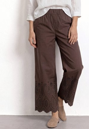 Femme portant un pantalon large marron avec un ourlet festonné brodé et des chaussures beige slip-on, debout contre un mur blanc texturé.