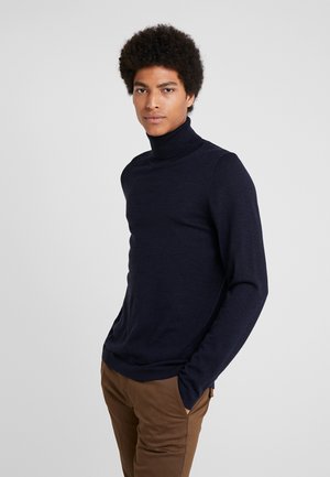 Marinblå polotröja i mjukt stickat material, med långa ärmar och en figurnära design. Modellen bär den med bruna byxor.