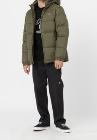 Oliwkowa puchowa kurtka z kapturem, elastycznymi mankietami i pikowaną fakturą. Noszona na czarnej bluzie i zestawiona z czarnymi spodniami oraz sportowymi butami.