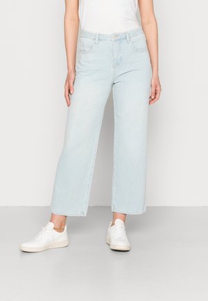 Slim-fit light blue cropped jeans worn with white sneakers on a standing person against a neutral background.