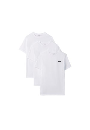 Jeune homme debout pieds nus portant un t-shirt blanc à manches courtes et un short bleu clair sur un fond gris uni.