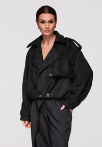 Femme aux cheveux foncés portant une veste noire double boutonnage oversize avec des boutons et un pantalon sombre sur mesure, posant devant un fond clair uni.