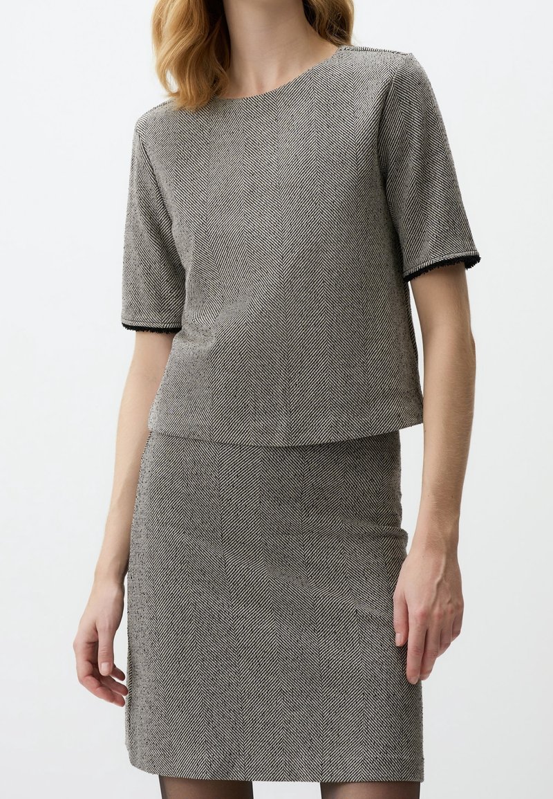Femme portant un haut gris à manches courtes avec un motif en chevrons et une jupe assortie sur un fond uni.