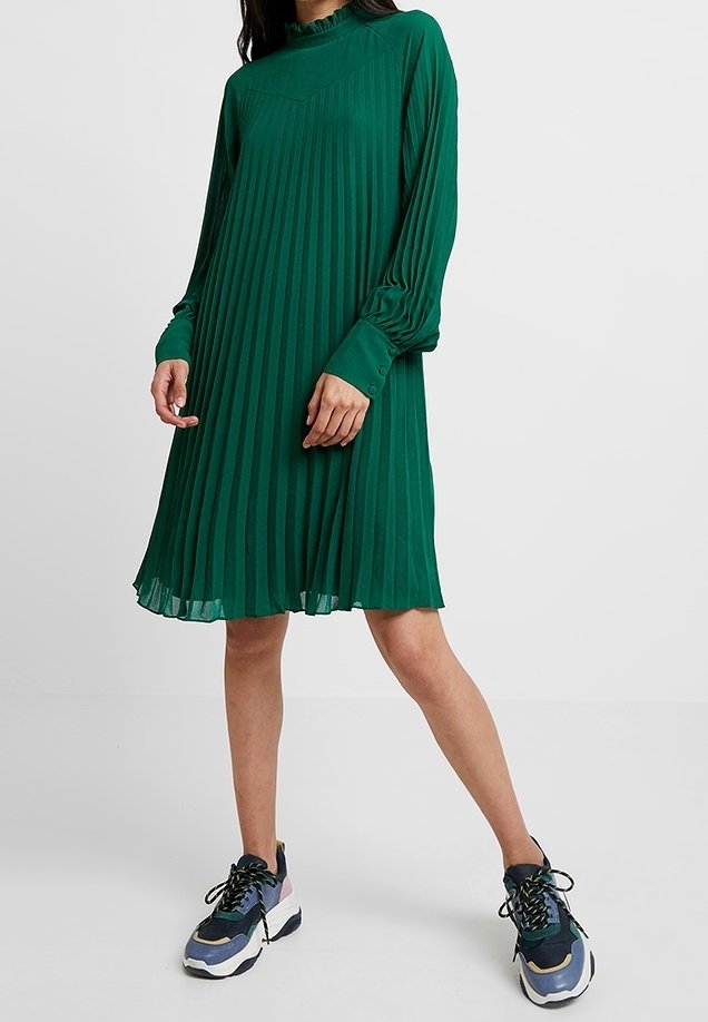 Vihreä pliseerattu mekko, jossa on pitkät hihat ja korkea kaula-aukko, valmistettu kevyestä kankaasta. Yhdistetty urheilullisiin tennareihin.