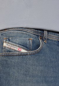 Blå denimjeans med en synlig ficka som har en vit etikett med texten "Diesel Industry, Denim Division". T-shirtens fåll synlig ovanför.