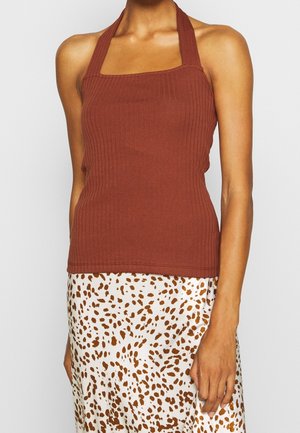 Femme portant un haut marron sans manches à col halter côtelé et une jupe blanche à taches abstraites marron, debout devant un fond uni.