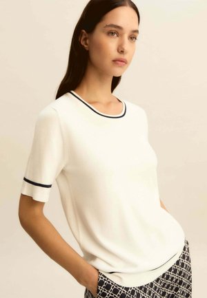 Donna con capelli lunghi e scuri che indossa una maglietta bianca a maniche corte con rifiniture nere e pantaloni a motivo bianco e nero, in piedi con le mani nelle tasche.