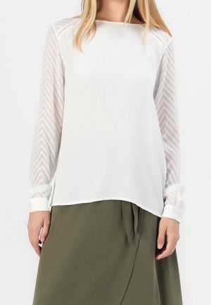 Woman wearing a white blouse with sheer patterned long sleeves and an olive green skirt with a front tie detail.