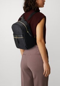 Sac à dos en nylon noir de forme arrondie, avec des zips dorés, une poche frontale et des bretelles réglables, porté sur un pull bordeaux et un pantalon beige.
