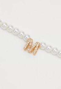 Bracelet en perles avec un charme en or en forme de lettre "M" incrusté de petits cristaux transparents sur un fond blanc.