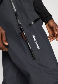 Fekete teljesítmény nadrág, stílusos dizájnnal, vízálló anyaggal, cipzáras zsebekkel és a "SALOMON" felirattal fehér színben a combon.