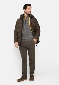 Mand iført brun dynejakke over grå kvart-lynlåssweater, olivengrønne bukser og brune sneakers, stående med den ene hånd i lommen.