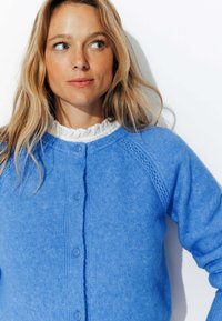 Cardigan bleu en matière tricotée douce, avec un col rond, une fermeture boutonnée et des manches raglan texturées.