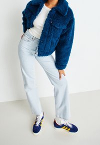 Blå fuskpälsjacka med två framfickor, ihop med ljusblå vida jeans och marinblå sneakers med gula ränder.