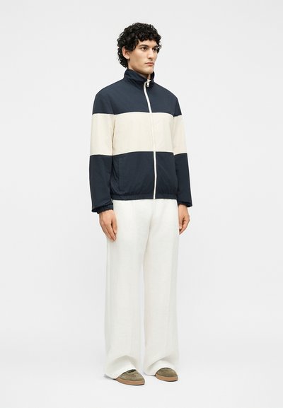 Άνδρας στέκεται φορώντας μαύρο-λευκό μπουφάν με φερμουάρ και μπλοκ χρωμάτων, φαρδιά λευκή παντελόνα και καφέ δερμάτινα παπούτσια σε απλό φόντο.