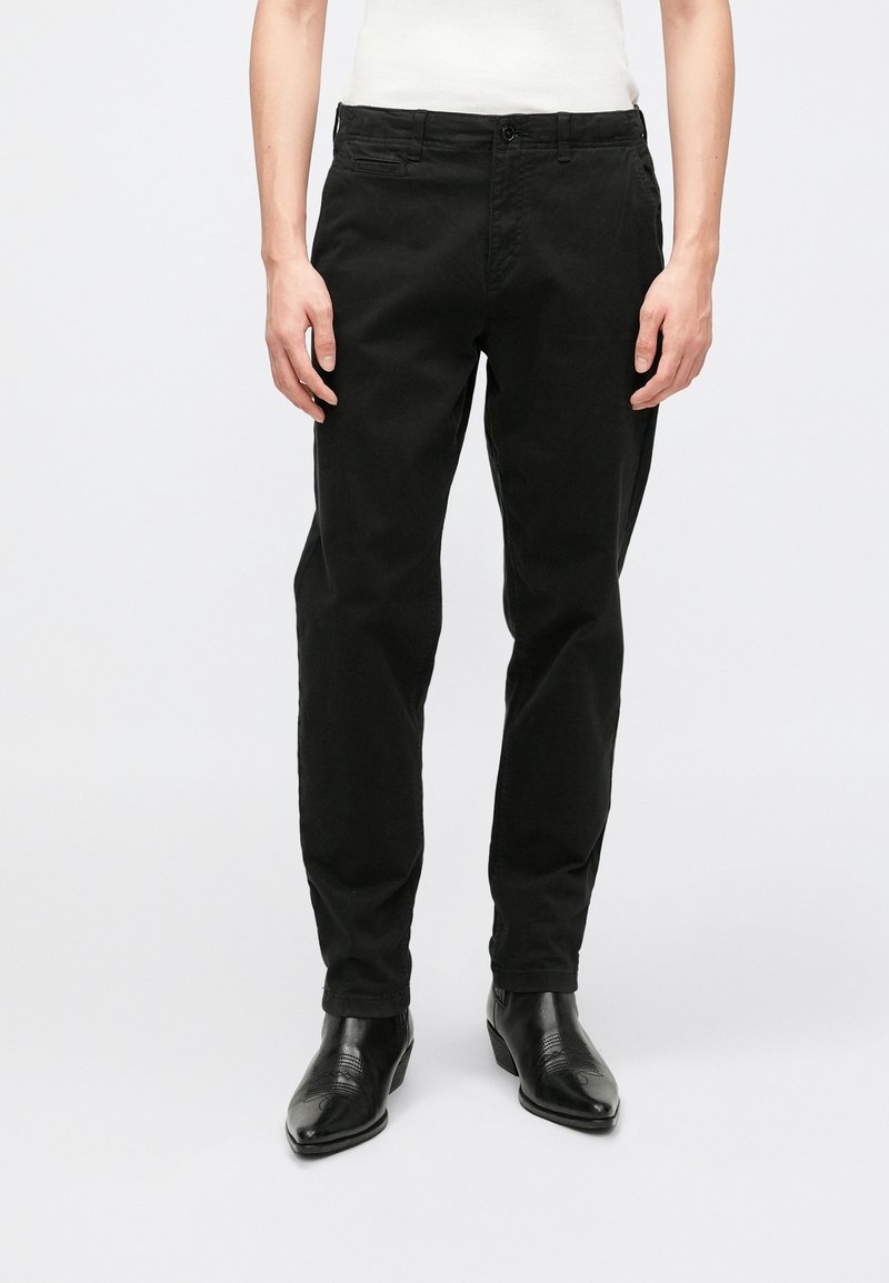 Homme portant un pantalon noir droit avec des poches avant, associé à des bottes en cuir noires à bout pointu, debout devant un fond uni.
