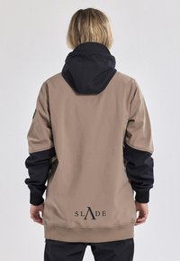 Beige und schwarze Kapuzenjacke mit glatter Textur, die ein Logo am Saum und kontrastierende Ärmel aufweist. Entspannt geschnittenes Design.