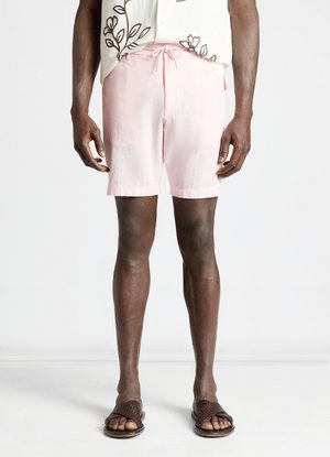 Homme portant un short à cordon rose clair, des sandales tressées marron et une chemise blanche avec une broderie florale marron, debout sur un sol blanc.