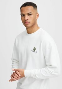 Balts kokvilnas džemperis ar garām piedurknēm, kurā kreisajā krūtī ir zaļš logs, apaļš kakla izgriezums un ribota aproce. Visā virsmā ir gluda tekstūra.