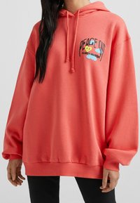 Sudadera de gran tamaño en color coral con capucha ajustable, puños de punto acanalado y un gráfico en la parte frontal que dice "PAZ FUERA" con un diseño de corazón y nubes.