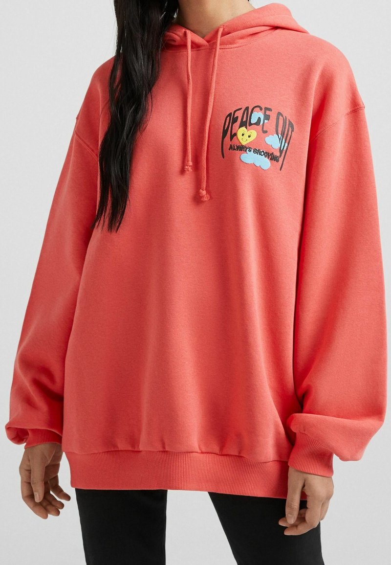 Sudadera de gran tamaño en color coral con capucha ajustable, puños de punto acanalado y un gráfico en la parte frontal que dice "PAZ FUERA" con un diseño de corazón y nubes.