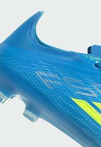 Chaussure de football synthétique bleue avec surface texturée, bandes jaune néon et crampons transparents sous la semelle, vue de côté.