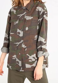 Kamouflagejacka i brunt, grönt och grått, med två bröstfickor, knappstängning och uppvikta ärmar. Tillverkad av slitstarkt tyg.