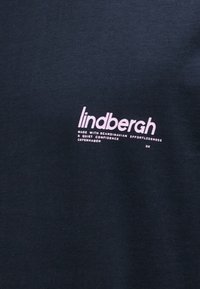 Μαύρο ύφασμα με ροζ κείμενο "lindbergh" και μικρότερο κείμενο που αναφέρει "Κατασκευασμένο με σκανδιναβική αβίαστη άνεση και ήσυχη αυτοπεποίθηση Κοπεγχάγη Δανία."