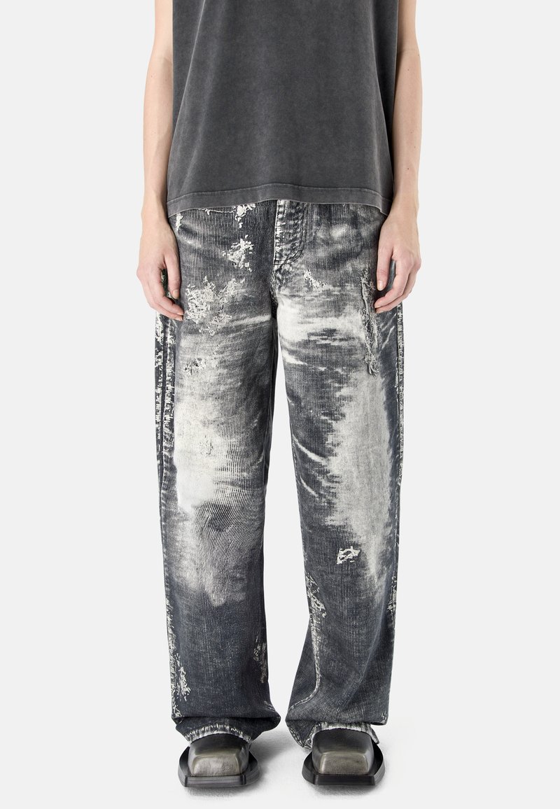 Personne portant un pantalon ample noir et gris délavé et usé, un haut noir sans manches et des chaussures noires à bout carré.