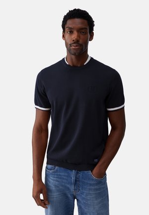 Uomo che indossa una camicia blu navy scuro a maniche corte con rifinitura bianca sulle maniche e sul colletto, abbinata a jeans blu, in piedi con una mano in tasca.