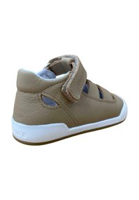 Zapatilla de piel marrón con suela de goma blanca, tira de velcro, detalle de corte en los laterales y un interior acolchado.